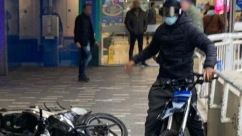 A blurry image of a man on a moped. He is wearing dark clothing and his face is covered with a mask and goggles. Another moped is lying on the floor next to him.