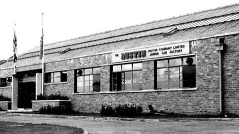 A black and white photo of the front entrance to a long bricked building. It shows the Austin logo, along with the Union flag, and another which is indistinguishable.