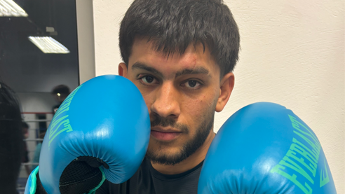 Shows young man with a dark hair and a beard in a dark T-shirt and light blue boxing gloves 