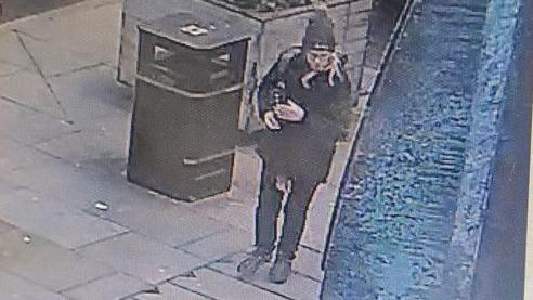 A CCTV image shows Ms Fish in a black coat and hat with black jeans and trainers walking past a rubbish bin outside a shop.