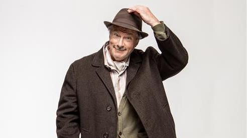 A man wearing a beige-coloured cardigan and a brown coat is lifting a hand up, holding onto a wide-brimmed brown hat.