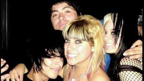 On the left is a boy with black hair. A girl with straight blonde hair and a lip piercing has her arm around him. A boy behind her wears a blue top and has short black hair. He has his arm around a girl to the far right wearing a black fish net top and has straight blonde and black hair. 