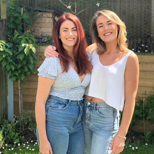 Leanne (left) and Sarah (right) smile as they stand in a sunny garden. Leanne has long red hair and Sarah has light shoulder length hair. 
