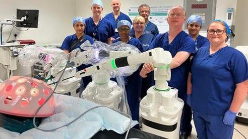 A team of surgeons wearing blue scrubs, hats and gloves stand in a theatre next to a large robotic arm. 