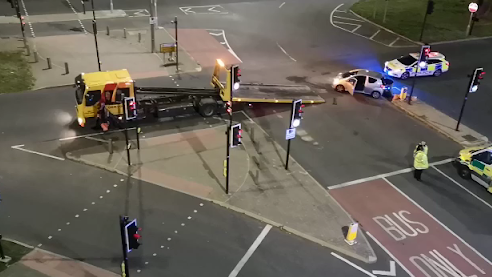 A car is crashed at the junction. A police car and ambulance are present, as is a truck and trailer, perhaps to carry the car away.