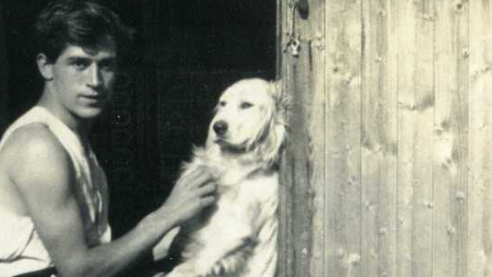 Black and white photo of David Brynley as a young man fussing a light-coated retriever-type dog. He has dark hair with a long fringe and is wearing a sleeveless vest.