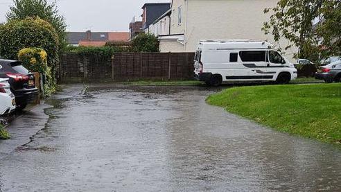 The image shows a residential street which is flooded with water and the rain is still coming down. You can see cars parked in a driveways to the left of the image and a camper van parked on the pavement to the right of the image. There is also a grass verge which to the right which is still visible above the water level.