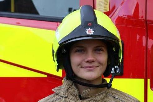 Phoebe Hutley is looking towards the camera smiling. She is wearing a yellow and black firefighter helmet that has an Essex County Fire & Rescue Service logo on the front. She is standing in front of a yellow and red fire engine.