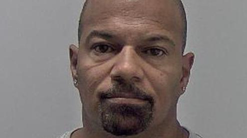 A mug shot of a man with a cropped black goatee, shaved head and diamond studs in both ears.