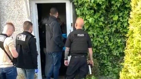 Four police officers can be seen entering the open door of a property, which has ivy growing down one side.