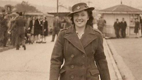 Phyllis Vincent pictured in a sepia image from World War Two, when she was a woman in her late teens or early 20s. She is smiling at the camera and wearing a Land Girl overcoat and brimmed hat. A group of schoolgirls and soldiers can be seen in the unknown background, which appears to be a seaside promenade.