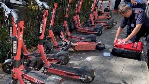 A man is kneeling down and opening a red bag. he is in front of a row of red e-scooters parked against a black park fence. Other people can be seen behind him.