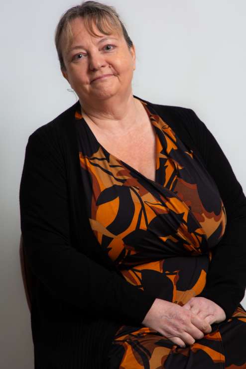 Woman in a leaf print brown black and orange dress. She has greying hair which is tied back.