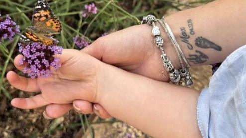 A small child's hand holds a purple flower with a butterfly on it. The child's hand is being supported by a woman's hand. She is wearing three silver bracelets and has a tattoo on the inside of her wrist of a baby's hand and foot next to the name Max and a date. 