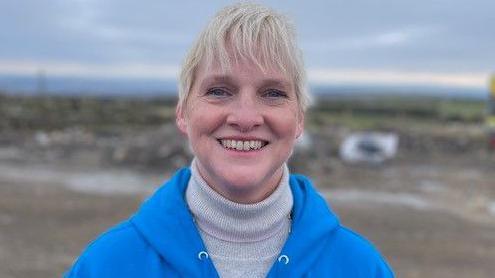 Amanda Briggs smiles at the camera. She is standing outside wearing a blue hooded top and light coloured turtleneck sweater. 