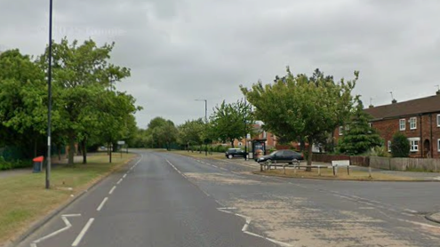 A street with houses on the left and trees on the right and a building with scaffolding on in the distance. There is another road going off to the right. It is a grey, cloudy day.