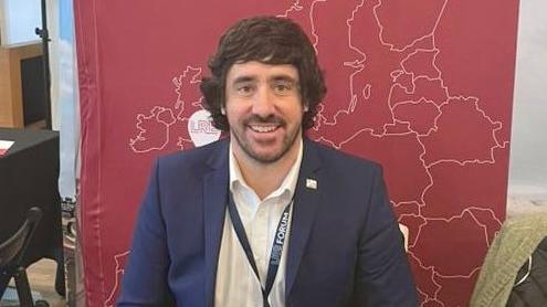 A man with brown hair and a beard, wearing a white shirt, dark blue jacket and lanyard, sits in front of a red background with a map of the Europe on it.