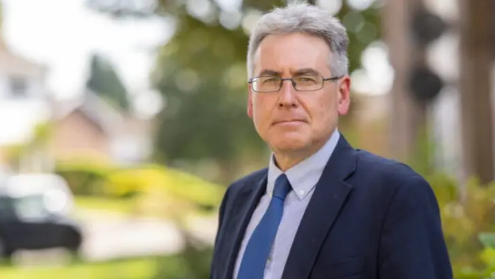 A man in a blue suit and tie with glasses is looking directly at the camera with a slight smile