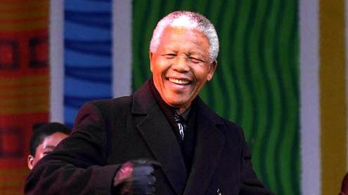 Nelson Mandela grins as he dances on stage at a concert in Leeds in 2001. There are singers behind him with a backdrop of green, blue and orange colours celebrating African culture.