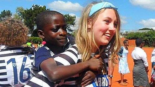 Katie in Malawi gives a piggy-back to a little boy during a football match. She is smiling broadly, wearing a football strip - other youngsters behind her are also wearing the strips and some girls stand in the background in the school playground.