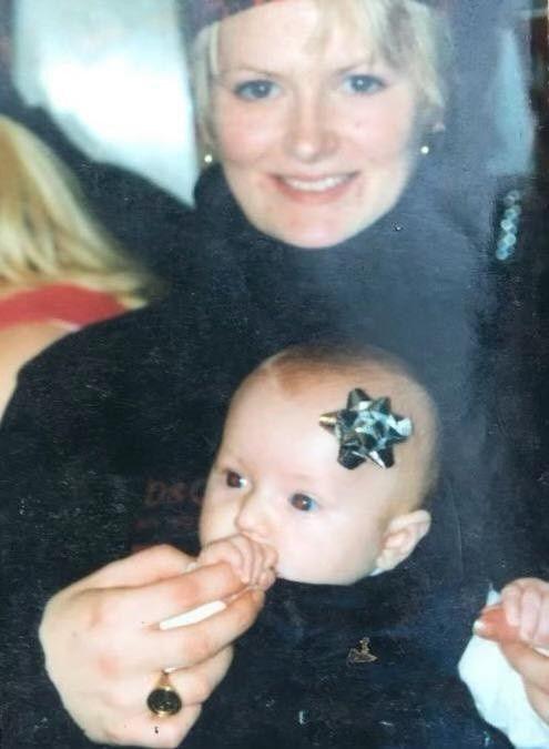 A woman holds a baby on her lap. The woman has short blonde hair, and she is wearing a black polo neck jumper and small gold hoop earrings. The baby is sucking on one of the woman's fingers and has a silver rosette on its forehead.