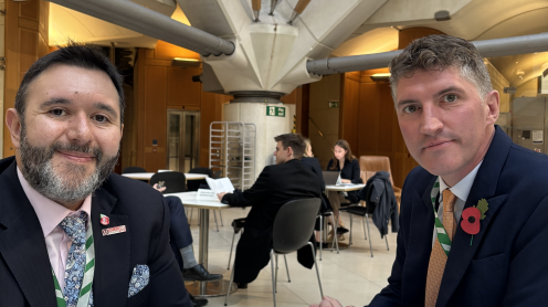 Two men are sat next to one another looking into the camera in what looks to be a lunch hall. They are dressed in black suits. Sadik has a lilac floral tie with a pink shirt, and Edward has a yellow tie with a white shirt. They both wear poppies.