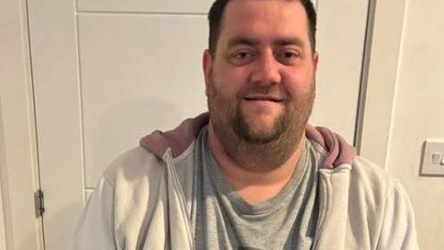 Patrick Murphy, situation in a house and smiling to camera. He has short brown hair and a short dark beard and is wearing a grey t-shirt and a white hoodie. 