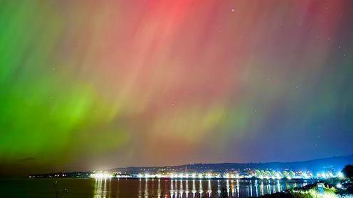 Green and red beams light up the night sky above the sea. A row of lights can be seen in the distance.