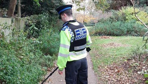 A young community support officer stands on the path of a park with a metal detector pointing at a bush.