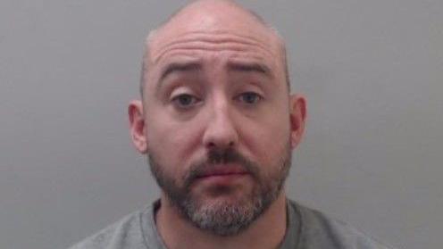 A man with a dark and grey beard looks into the camera. He is bald and is wearing a grey sweatshirt.