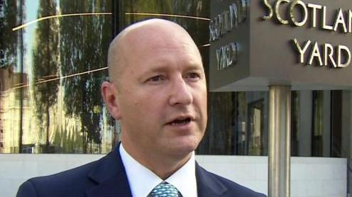Commander Dominic Murphy is standing outside the Met Police headquarters in London. A sign behind him says "Scotland Yard". He is balding and is wearing a blue suit jacket, a white shirt and a patterned light blue tie