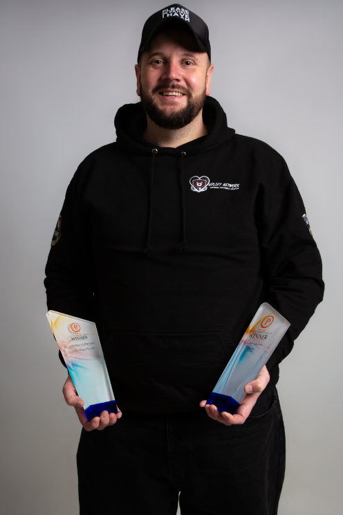 Man with beard wearing a black cap. He's holding two awards.
