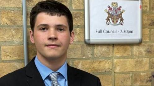 Hayden with short dark hair looking at the camera while wearing a black jacket, blue shirt and grey tie. He is standing in front of a brick wall on which there is a notice with the council crest and "Full Council - 7.30pm" in black lettering. There is a wooden door to the right with a large blue and white sticker indicating that the door is automatic and there is a hearing loop installed.
