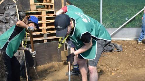 A man and a woman both wearing dark green St Andrew's Healthcare jackets and caps, dig holes with spades in the earth. 