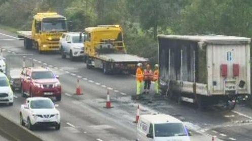 A CCTV image showing cars passing by the burnt load via two lanes. The burnt load is cordoned off with three people in high vis standing next to it.