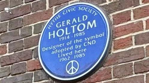 A circular blue plaque reading "Hythe Civic Society / Gerald Holtom / 1914 -1985 / Designer of the symbol adopted by the CND / Lived here / 1962 -1985"