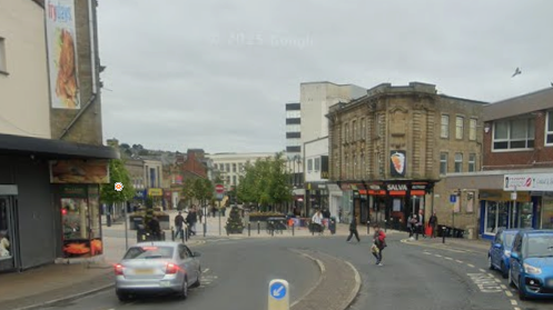 Image shows a Google Streetview shot of Yorkshire Street in Burnley. It is a section of road that ends with the beginning of a pedestrianised area, which is lined with shops and fast food restaurants.