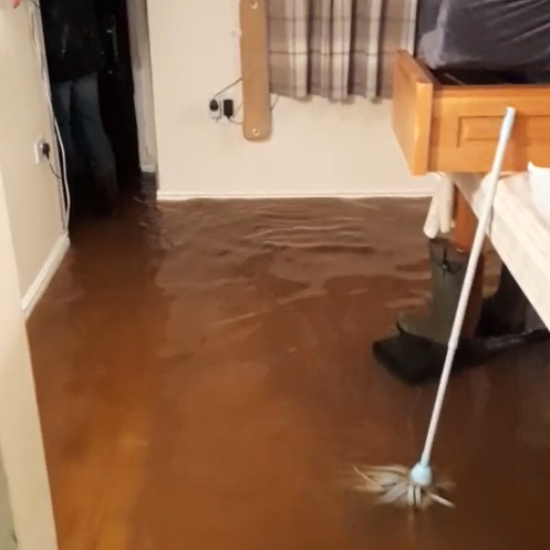 A photo taken by Paul during the flooding - the wooden floor is submerged in brown, discoloured water. A mop stands leaning against a dining table, which is covered in various belongings picked up out of the water. 
