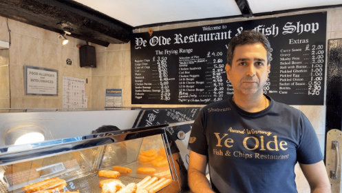 Shahram Navard looks into the camera with a solemn expression. He is wearing navy t-shirt with 'Ye Old Fish and Chips Restaurant' on his t-shirt. He is leaning against the counter where the chips and food are visible. Behind his is a blackboard with prices written on in chalk. 