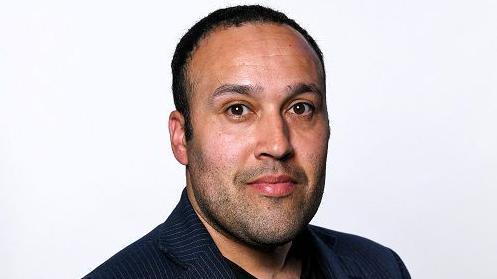 Barry Crisp with short dark hair and slight beard. He is wearing a dark jacket and is in front of a white background.