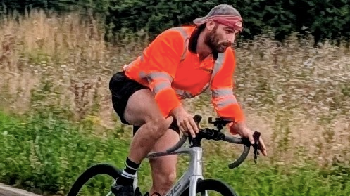 Kieran Palmer riding a bike outside. He is focusing on the road in front of him and is not looking at the camera. He is wearing black shorts, a baseball cap and a long-sleeve orange T-shirt with fluorescent stripes on it.