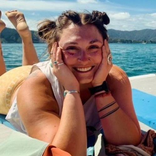 Emilie Anne Parker looking directly at the camera with her hands on either side of her face while propped up on her elbows. She is wearing a yellow skirt with a white top and has her feet raised behind her in front of a body of water with mountains and blue sky in the background.