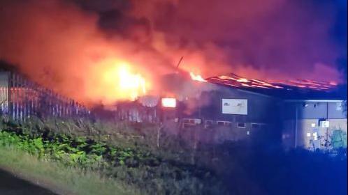 Flames ripping through the roof of warehouse buildings at night, with grass in the foreground.