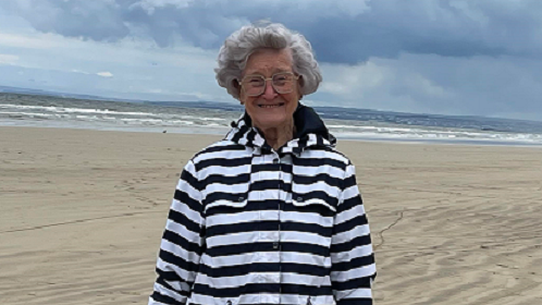 Joyce Taggart is smiling whilst wearing a black and white striped coat with neutral trousers. She is standing on the beach.