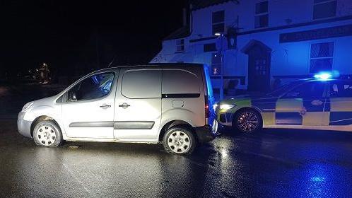 A silver van with flat tyres next to a police car with its lights on. In the background is a building illuminated by blue police lights. The sky is dark.