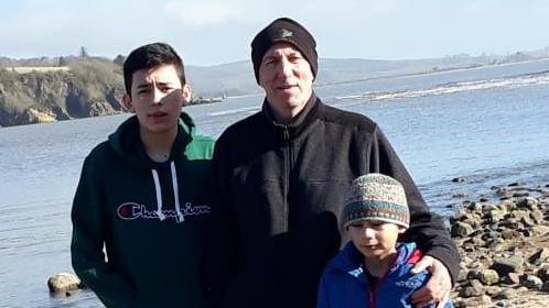 A man in a black hat and black top and jeans stands with two children - one in a green hooded top and the other in a blue anorak in front of the rocky Dumfries and Galloway shoreline