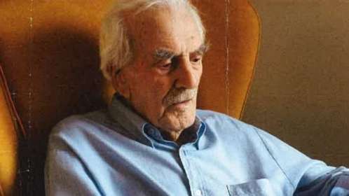 Mr Monk is sat in an armchair with a blue shirt on holding what looks like an ipad or an item of technology. He has grey hair and  moustache and the chair is orange. 