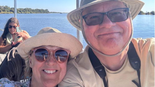 The couple have taken a close-up selfie and both a smiling and wearing hates as they travel on a boat on the river.