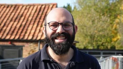 Evangelos Achilleos smiles at the camera as he stands outside. He is bald and has a dark, thick beard. He has glasses and wears a blue polo top.