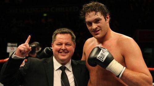 Fran's father, Mick Hennessy, wearing a suit, standing in a boxing ring with boxer Tyson Fury, who holds up a fist with a boxing glove on.